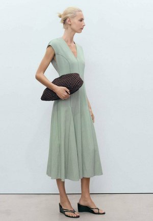 Robe sans manches vert clair avec un décolleté en V profond et une jupe évasée. Associée à une pochette texturée marron et des sandales en tongs noires.
