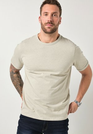 Uomo con capelli corti e barba, che indossa una t-shirt beige chiaro, jeans scuri e un orologio, in piedi con le mani dietro la schiena contro uno sfondo bianco.