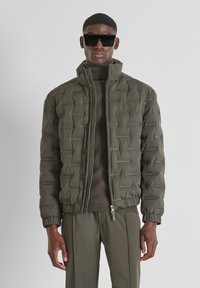 Giacca puff verde oliva con texture trapuntata, colletto alto e chiusura con zip. Indossata sopra un maglione a costine abbinato e pantaloni aderenti.