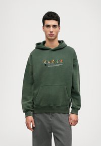 Zaļš džemperis ar kangaru kabatu, kapuci ar šņorēm un izšūtu daudzkrāsainu dizainu, kas attēlo multfilmu figūras visā krūtīs.