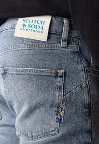 Lichtblauwe denim jeans met een wit label met blauwe tekst. De achterzak heeft decoratieve stiksels in blauw, geel en bruin.