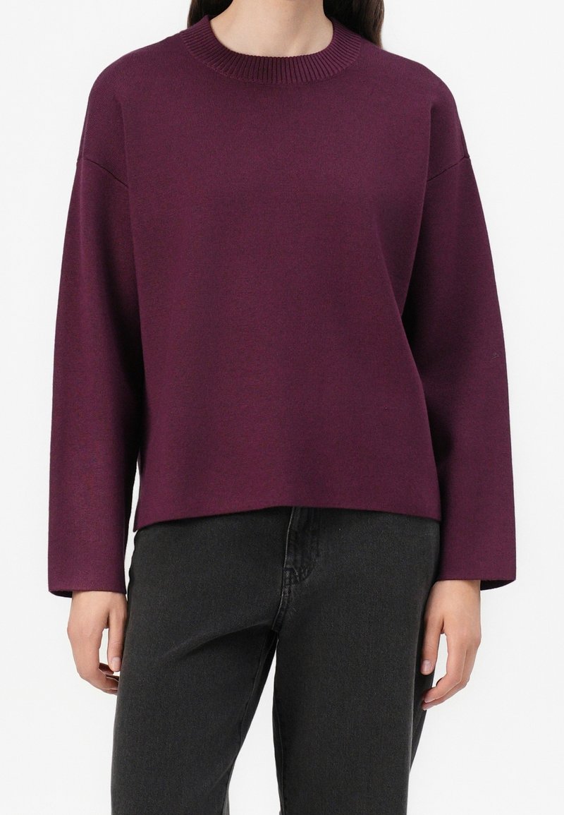 Femme portant un pull violet foncé à manches longues et col rond, associé à un jean noir taille haute sur un fond uni.