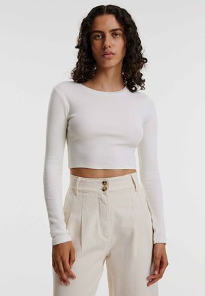 Weißes, geripptes Langarm-Crop-Top mit rundem Ausschnitt, kombiniert mit hoch taillierten cremfarbenen Hosen mit zweifach Knopfverschluss auf der Vorderseite.