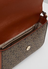 Hnědá monogramová crossbody taška se zlatým zapínáním na knoflík, otevřená, ukazující hladkou hnědou koženou klopu a béžový látkový interiér s malou kapsičkou.