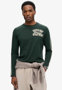 Mørkegrøn langærmet skjorte med "SUPERDRY Athletic Department" i cremefarvet tekst. Har hvide sidelister, blød tekstur og en rund halsudskæring.