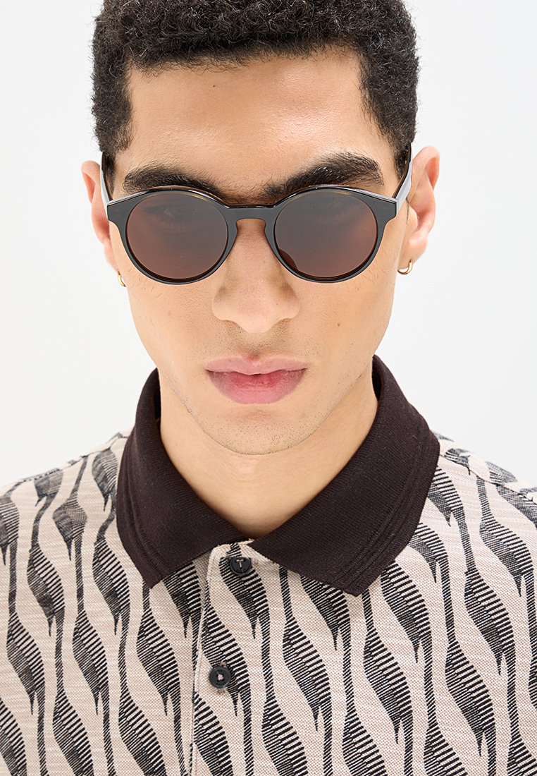 Runde Sonnenbrillen mit dunklen Gläsern und schwarzem Rahmen. Das Modell trägt ein gemustertes Polohemd in Beige und Schwarz mit einem dunklen Kragen.