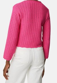 Pull en tricot rose avec un motif texturé, manches longues et coupe courte. Porté avec un pantalon blanc taille haute, avec poches arrière.
