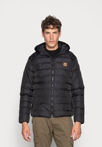 Black puffer jacket with a hood, featuring horizontal quilted design, front zipper, and a tan logo patch on the left chest area.