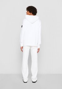 Felpa bianca con cappuccio regolabile, polsini a costine e una patch sulla manica sinistra. Abbinata a pantaloni bianchi a gamba larga e scarpe nere.