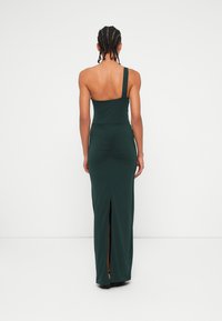 Γυναίκα με πλεξο�ύδες που φοράει ένα σκούρο πράσινο φόρεμα maxi με ράψιμο ενός ώμου και άνοιγμα στην πλάτη, στέκεται μπροστά σε έναν απλό λευκό φόντο.