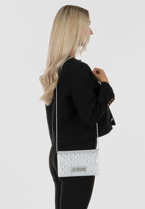 Borsa a tracolla in argento con motivo a trapunta, dotata di una catena e una piastra con logo, di forma rettangolare. Indossata contro un giubbotto scuro, vista laterale.