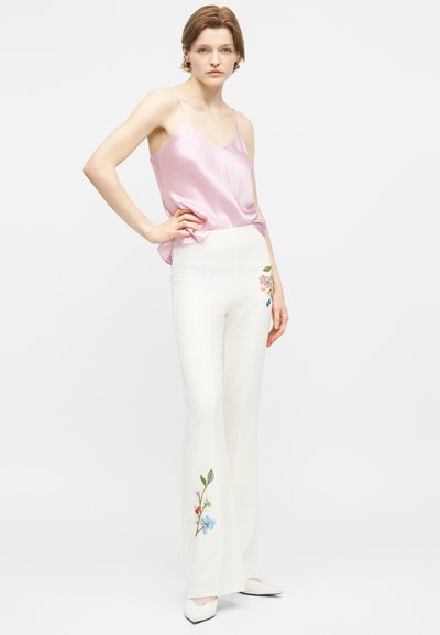 Γυναίκα που φοράει λαμπερό ροζ καμιζόλ και λευκό ψηλόμεσο παντελόνι με πολύχρωμη κεντητή λουλουδένια διακόσμηση, στέκεται με το χέρι στη μέση.