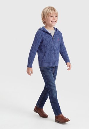 Blond chłopiec idzie, ubrany w niebieską sweterkową bluzę zapinaną na suwak, szarą koszulkę, ciemnoniebieskie jeansy i brązowe skórzane botki za kostkę, uśmiechając się.