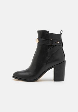 Bottine en cuir noir avec talon bloc, bout pointu et sangle décorative avec boucle dorée sur le côté.