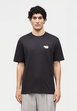 Mandlig model med kort krøllet hår iført en sort t-shirt med et lille hvidt "N°7" logo på brystet og lysegrå bukser.