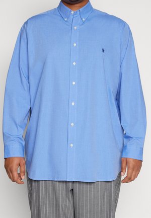 Mand iført en lyseblå, knappet, langærmet skjorte med et lille mørkt logo på venstre bryst og grå bukser med smalle striber.