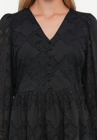 Schwarzes Kleid mit Spitzenblumenmuster, Knopfverschluss vorne, V-Ausschnitt und langen Puffärmeln, gezeigt von Kragen bis Taille an einer Person.