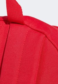 Esquina de una mochila de tela roja que muestra el material texturizado, costuras visibles y una lengüeta de tela. El diseño es compacto y funcional.