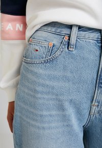 Los jeans de mezclilla azul claro presentan un diseño clásico de cinco bolsillos con herrajes en tono cobre, un pequeño parche con el logo y una textura ligeramente desteñida.