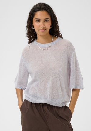Frau mit lockigem dunklem Haar trägt einen hellvioletten durchsichtigen Kurzarmpullover, braune Hose, silberne Halskette und Creolen, Hände in den Taschen.