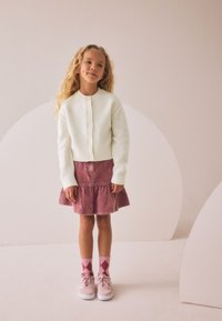 Balta adīta kardigana, rozā korduiro auduma svārki ar volānu apakšmalu, rozā zeķes ar dimanta rakstu un gaiši rozā sporta apavi uz neitrāla fona.