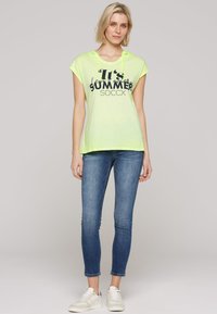 Neon yellow hooded t-shirt with black printed text, paired with fitted blue denim jeans and white sneakers. Casual summer outfit.