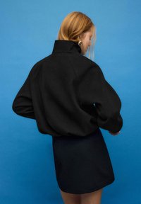Sort oversizejakke med høj krave og brede ærmer, kombineret med en kort sort nederdel, foran en blå baggrund. Glat stofstruktur.
