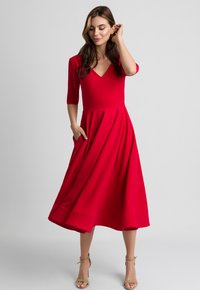 Kobieta w czerwonej sukience midi z dekoltem w serek, o długości rękawów do trzech czwartek, w beżowych sandałach na kostkę, stojąca na tle jasnoszarym.