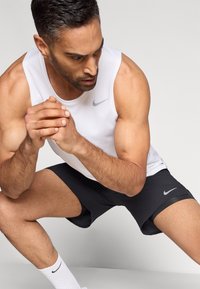 Άνδρας με λευκή αθλητική μπλούζα χωρίς μανίκια και μαύρο σορτς τεντώνει τα πόδια του με τα χέρια δεμένα, κοιτάζοντας συγκεντρωμένος σε φόντο απλό.