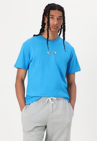 Modra bombažna majica s krožnim ovratnikom in kratkimi rokavi, z srebrnim logotipom na sredini. Kombinirana z sivo jogger hlačami.