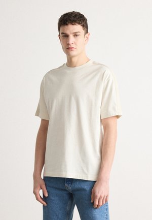 Beige t-shirt med kort ärm i bomull. Har rund hals och avslappnad passform. Matchas med ljusblå denimjeans.