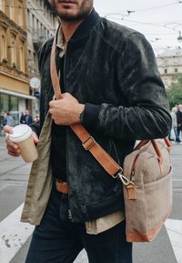 Brown canvas bag with leather accents, shoulder strap, and zipper. Black suede jacket with ribbed cuffs over a tan shirt. Coffee cup held in hand.
