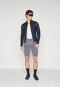 Roupa de ciclismo composta por um casaco azul-marinho ajustado, uma camada base branca e calções cinzentos. Inclui sapatos de ciclismo pretos e óculos de sol. Textura suave.