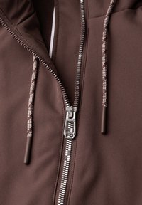 Fermeture éclair en métal partiellement fermée sur un sweat à capuche marron avec des cordons à motifs et une texture de tissu lisse.