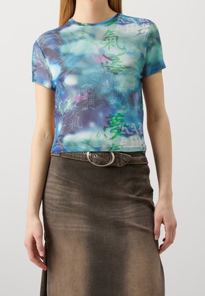 Kobieta ubrana w niebiesko-zielony top z krótkim rękawem w stylu tie-dye z azjatyckimi znakami, zestawiony z wyblakłą brązową spódnicą z wysokim stanem i srebrnym paskiem z klamrą.