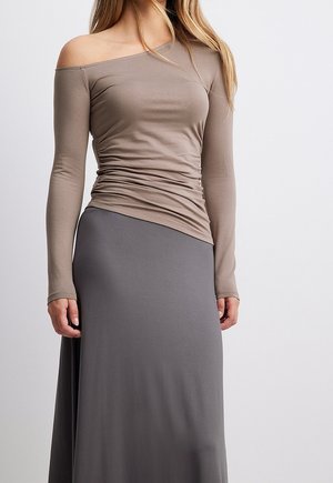 Femme portant un haut taupe à manches longues et épaules dénudées avec des côtés froncés et une jupe longue taille haute gris foncé sur un fond uni.