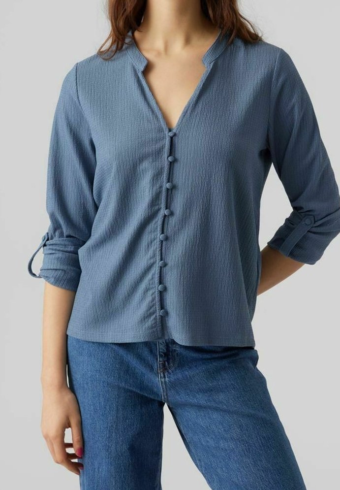 Femme portant un chemisier bleu texturé à manches longues avec des boutons recouverts de tissu et des manches retroussées, associé à un jean en denim bleu.