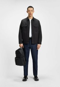 Sort jakke med brystlommer over en hvid skjorte, parret med mørkeblå denimbukser. Sort rygsæk holdt i hånden. Sorte sko.