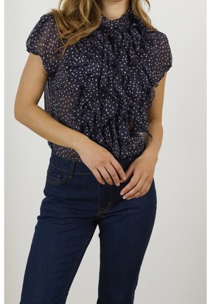 Marineblouse van doorschijnende stof, met een gerimpelde voorkant en een stippenpatroon. Gecombineerd met donkere spijkerbroek en handen op de heupen.