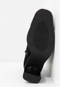 Chaussure en suédine noire avec un bout rond, un talon de hauteur moyenne, une semelle en caoutchouc texturé et des détails en métal argenté à l'arrière.