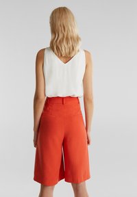 Bílý bezrukávový top s výstřihem do V spárovaný s vysokými oranžovými kalhotami culottes s bočními kapsami. Hladká látka s mírným leskem.
