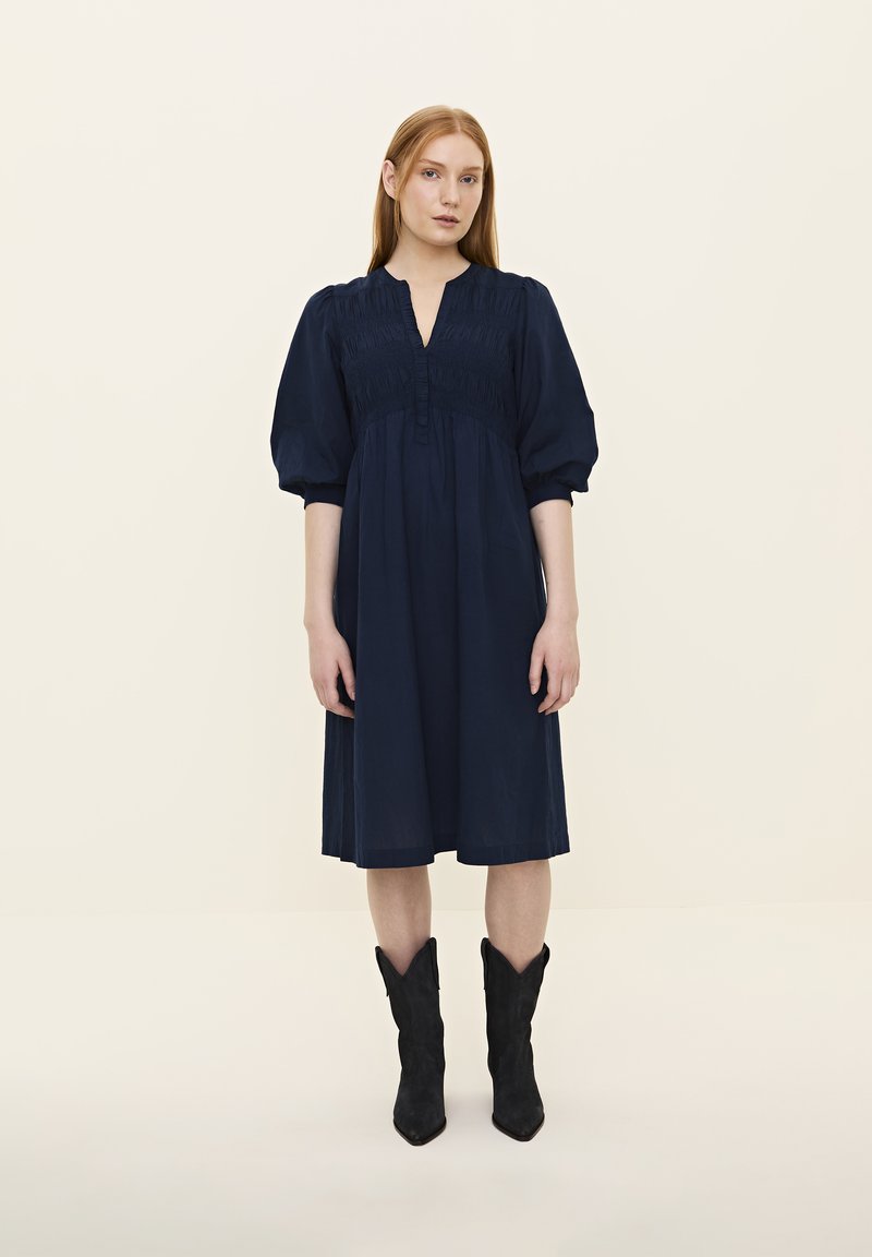 Marineblaue knielange Kleid mit V-Ausschnitt, gerafftem Oberteil und kurzen Puffärmeln. Kombiniert mit schwarzen Cowboy-Stiefeln.