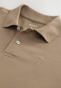 Camisa polo de color marrón claro exhibida de forma plana, con cuello, dos botones blancos y un pequeño logotipo bordado en el pecho. Marca visible en el interior.