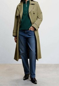 Olivgroene trenchcoat met een enkele knoop, gecombineerd met een donkergroen sweater met rits en een donkere blauwe spijkerbroek, zwarte schoenen met hak.