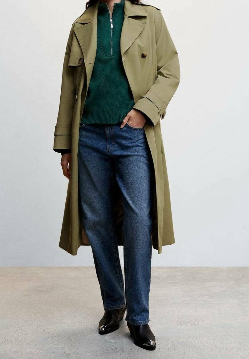 Olivgroene trenchcoat met een enkele knoop, gecombineerd met een donkergroen sweater met rits en een donkere blauwe spijkerbroek, zwarte schoenen met hak.