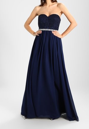 Granatowa wieczorowa suknia bez ramiączek z koronkowym gorsetem, biżuteryjnym paskiem i zwiewną spódnicą, noszona przez kobietę z rękami na biodrach.