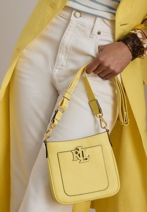 Osoba v bílých kalhotách a žlutém kabátu drží malou žlutou koženou kabelku s logem RL a širokým páskem, doplněnou náramky.