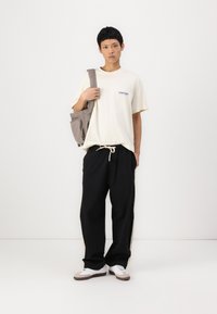 Lys beige T-shirt med lille blåt logo, sorte breddebukser med sidelister og hvide sneakers med grå accenter. Enkelt, casual outfit.