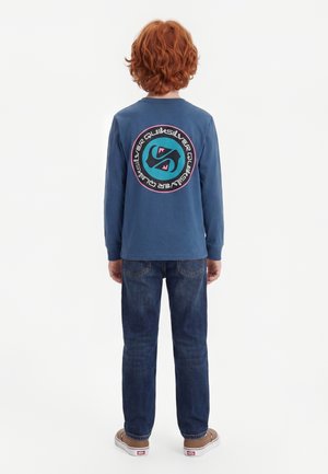Barn med rødt krøllet hår står med ryggen til, iført en blå langærmet trøje med en rund Quiksilver-logo på ryggen, mørke jeans og brune sko.