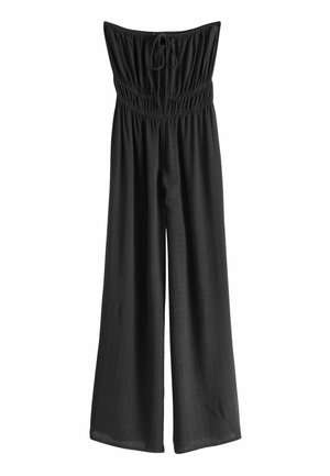 Femme portant une combinaison noire bustier et des sandales noires, tenant un sac fourre-tout tissé sur un balcon carrelé avec une rambarde bleue.